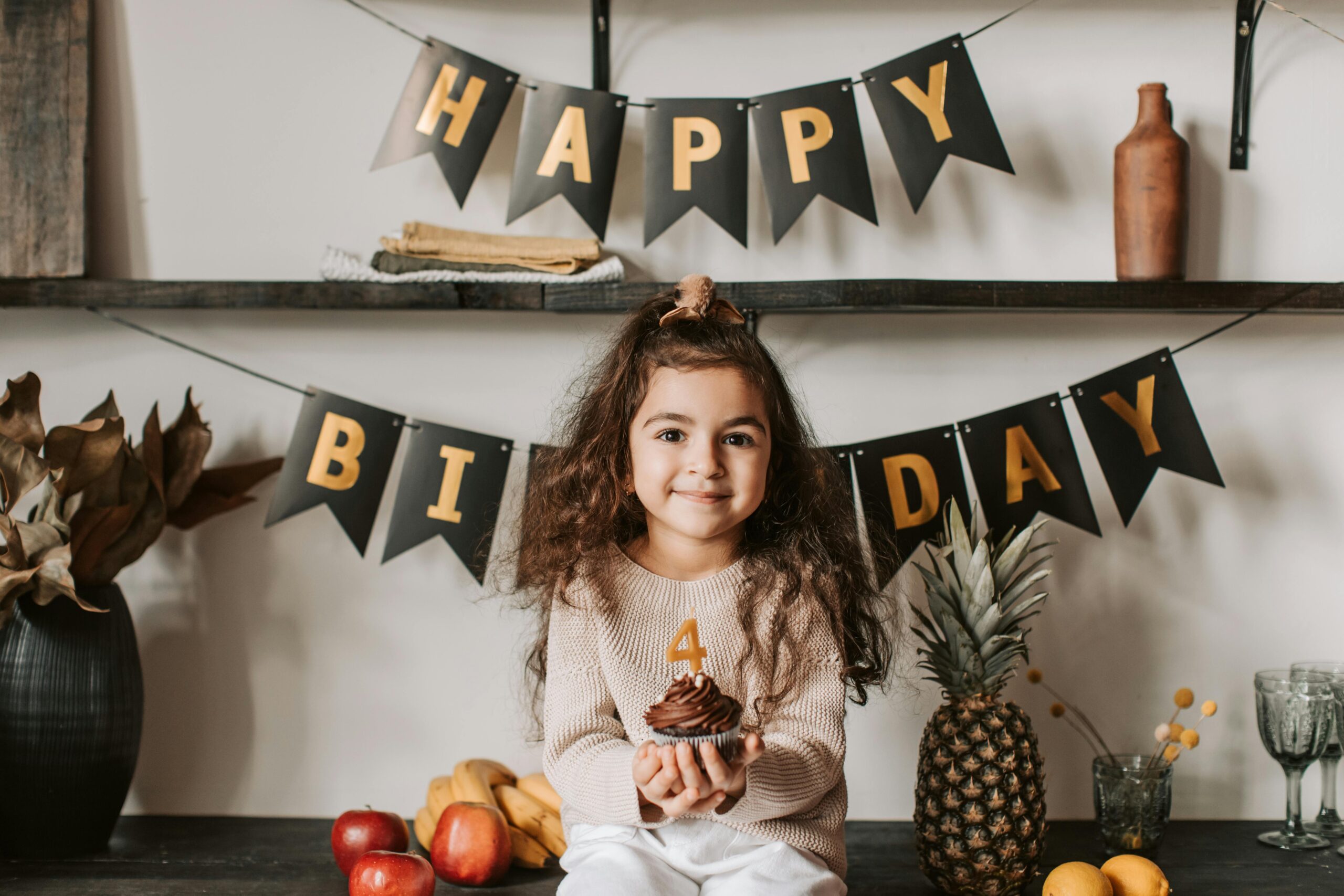 A smiling girl celebrates her birthday with a chocolate cupcake and fruit decorations indoors.